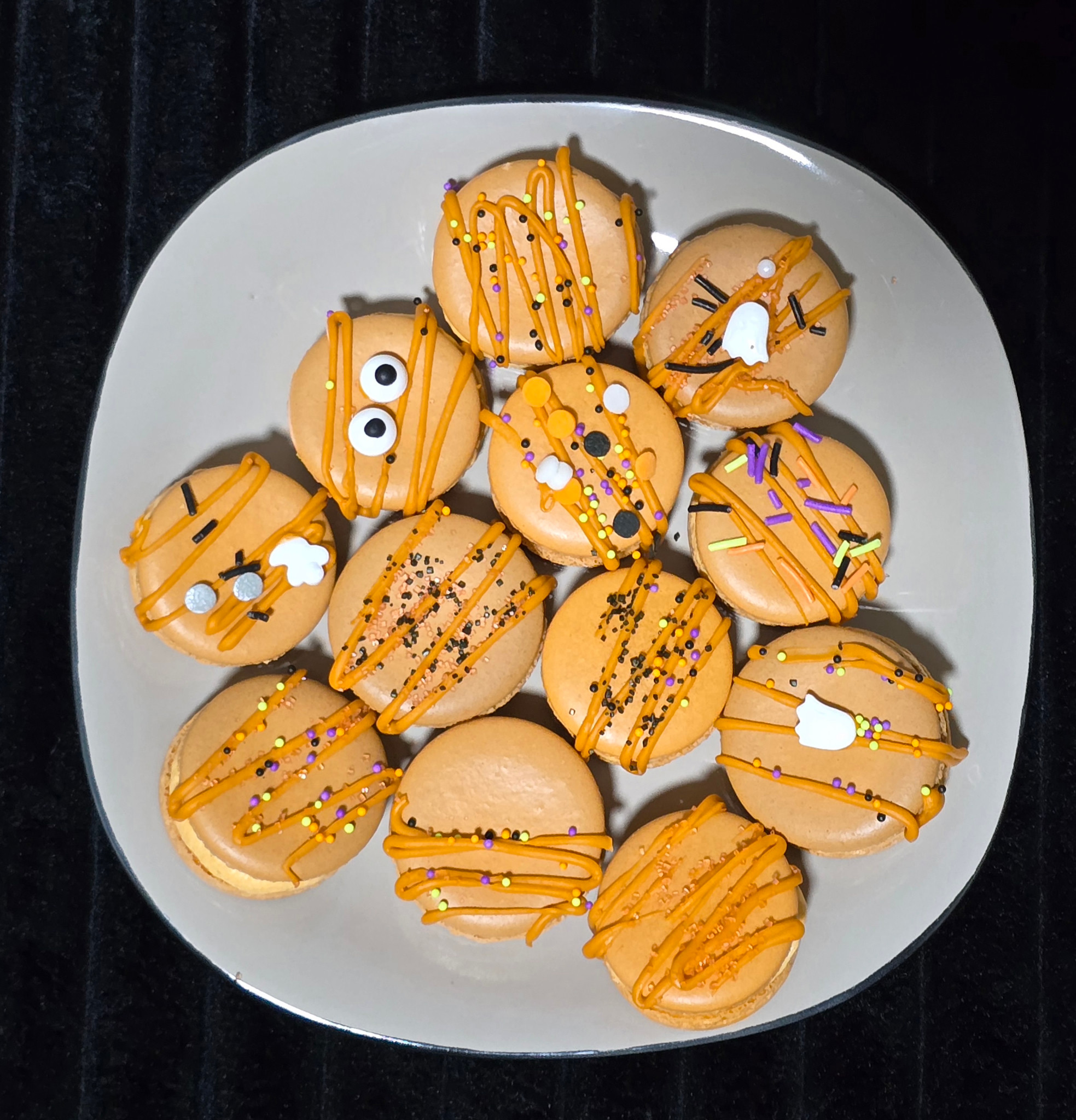 Pumpkin spice macarons on a white plate, decorated with orange drizzle, colorful sprinkles, candy eyes, and white ghosts.