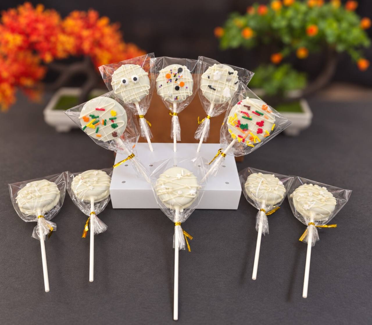 Individually wrapped dipped cookie pops with white drizzle, autumn sprinkles and candy eyes, arranged on a white stand.