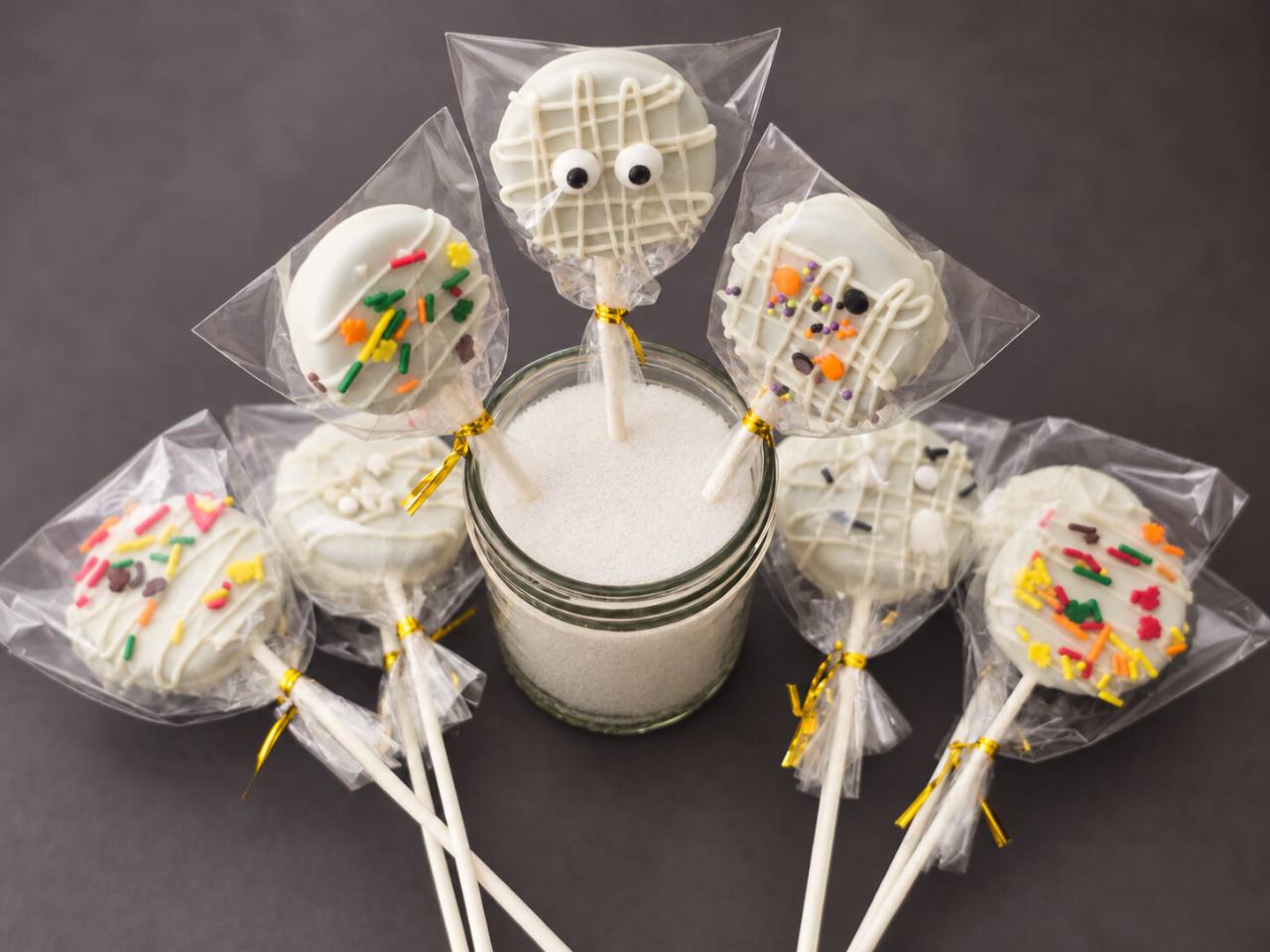 Three decorated cookie pops standing in a glass jar, surrounded by more wrapped pops.